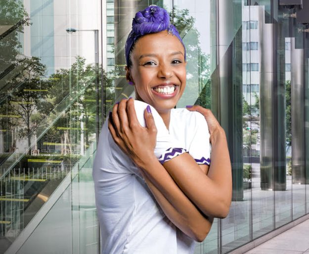 Embrace Equity A photograph of a woman embracing herself with her arms wrapped across her body. She has purple hair and is smiling towards camera.