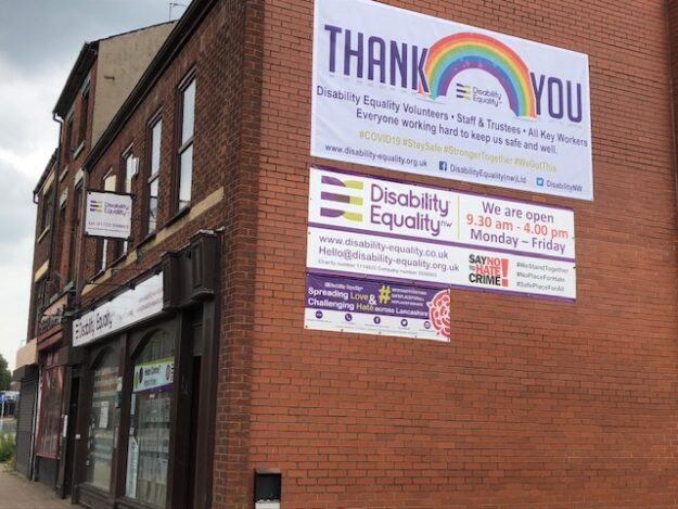 IMG_4351 Photo of side shot of the DENW building on Church street in Preston, with banners on the side of the wall including a Thank you NSH rainbow, DENW opening times and a Hate Crime logo. The text is blury in the image to see any further text.