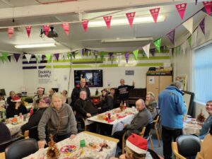 Photograph of our Festive Friday held in December of 2023. Lots of people are sat around tables that have Christmas tablecloths and decorations on the tables. Some of the people are wearing festive hats etc.