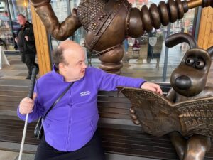 Photo of David sitting on the Wallace and Gromit bench in Preston, wearing a purple DENW branded fleece. 