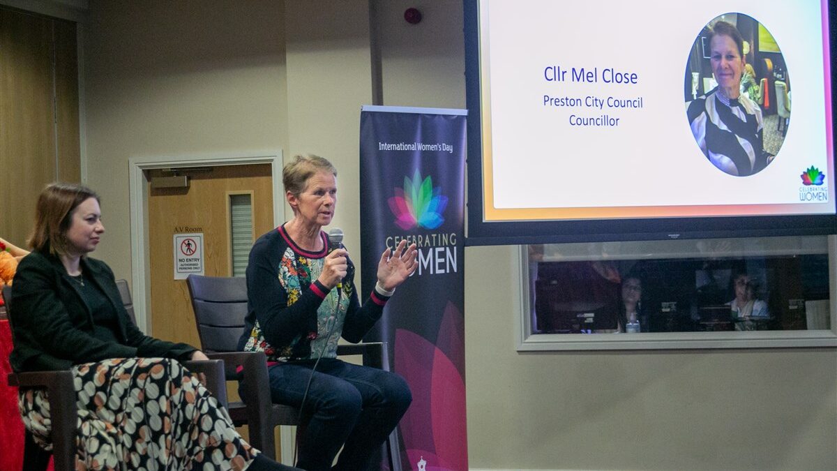Photo of CEO Mel Close at the BPS centre in Preston. Mel is wearing a black top with a colourful pattern on, holding a microphone on a stage alongside a fellow panelist. To the right there is a screen with Mel's name and headshot on.