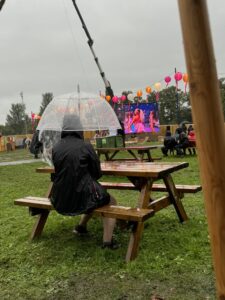 Wide shot of someone facing away from the camera, sat on a 4-seater bench, sat holding a clear brolly over their head.