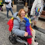 Person in a wheelchair waving a pride flag, on the main street in Preston, also wearing a rainbow feather boa and smiling at the camera.