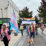 Wideshot of the Preston main street as the parade comes towards the camera. Someone holding a trans flag to the side and a cute dog at the front of the camera wearing a rainbow neckerchief.