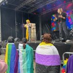 Further live shot of the BSL Interpreter, including people wrapped in pride and Trans flags stood at the front of the barriers to the stage.