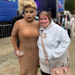Photo of Drag Queen Good Golly Miss Molly in peach sequin dress, and lady with grey hoody, pink handbag and face paints, stood in front of the PRIDE stage.