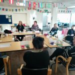 Wide shot of group with lead trainer at the front of the room, all sat in the Community Hub large room.