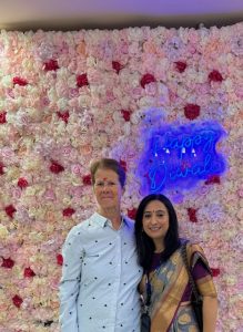 Mel Close stood next to a lady in traditional dress with a flower wall behind them and a neon sign saying "Happy Diwali"